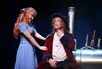 Siniseen mekkoon pukeutunut nainen hymyilee ja koskettaa punasamettitakkiin ja mustaan hattuun pukeutuneen vanhemman miehen olkapäätä, joka istuu kiiltävän hopeisen laitteen vieressä näyttämöllä, jossa on teatterivalot.