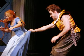 Keltaiseen ja ruskeaan pukuun pukeutunut mies tarttuu lavalla olevan siniseen pukuun pukeutuneen punatukkaisen naisen mekkoon, ja molemmat näyttävät olevan keskellä dramaattista kohtausta.