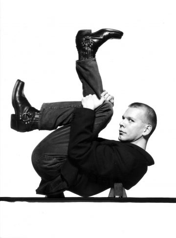 Pukuun ja farkkuihin pukeutunut henkilö makaa selällään, jalat taivutettuina rintakehän yläpuolella, pitää jaloistaan kiinni ja katsoo kameraan valkoista taustaa vasten.