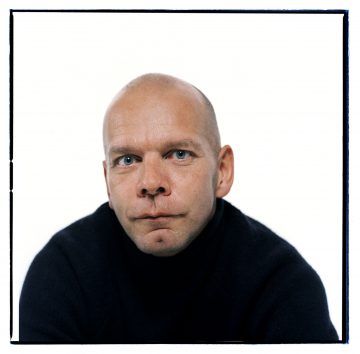 Kalju mies, jolla on siniset silmät ja vaalea iho, päällään musta villapaita, katsoo suoraan kameraan valkoista taustaa vasten.