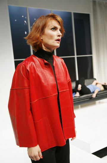 Lyhyet punaiset hiukset ja kirkkaanpunainen takki on lyhyellä punaisella naisella, joka seisoo etualalla ja näyttää vakavalta. Hänen takanaan toinen henkilö löhöilee mustalla sohvalla modernissa, minimalistisessa huoneessa, jossa on suuret ikkunat.