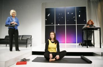 Kolme naista on modernissa lavastuksessa: yksi sinipukuinen seisoo mustan sohvan vieressä, yksi kelta-mustapukuinen istuu ristissä lattialla ja yksi harmaapukuinen polvistuu pöydän ääressä, kaikki suuren ikkunan edessä, jossa on violetti taivas.