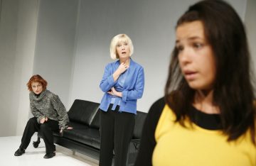 Lavalla on kolme naista: yksi punatukkainen nainen kyyristyy mustalla sohvalla, vaalea nainen sinisessä takissa seisoo yllättyneenä, ja ruskeaverikkö keltaisessa villapaidassa on etualalla huolestuneen näköisenä.