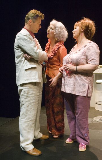 Lavalla seisoo kolme ihmistä: vaaleaan pukuun pukeutunut mies kädet ristissä, hänelle puhuu oranssiin puseroon pukeutunut vanhempi nainen, ja toinen vaaleanpunaiseen asuun pukeutunut nainen katselee heitä. He näyttävät käyvän dramaattista keskustelua.