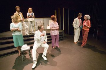 Seitsemän näyttelijää seisoo ja istuu lavasteissa värikkäissä puvuissa, osa valkoisissa puvuissa ja osa rennoissa vaatteissa, ja keskustelevat osana teatteriesitystä. Tausta on tumma ja lavasteet ovat minimaalisia.