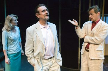 Lavalla kolme näyttelijää, kaksi miestä ja yksi nainen, kaikki vaaleisiin pukuihin pukeutuneita. Nainen näyttää huolestuneelta, toinen mies elehtii kädellään, ja toinen seisoo kädet taskussaan mietteliään näköisenä.