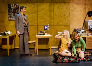 Pukumies seisoo kahden pöydän vieressä, ja kaksi naista istuu lattialla värikkäällä matolla. Toinen nainen juo kupista, ja toinen katsoo mattoa. Ympäristö muistuttaa vanhaa toimistoa.