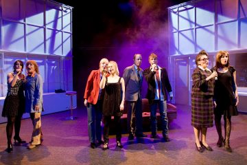 Kahdeksan ihmistä seisoo lavalla, jokaisella on mikrofoni kädessään ja he laulavat. Värikäs näyttämövalaistus ja savu luovat dramaattisen tunnelman, ja taustalla näkyy moderneja lavasteita.