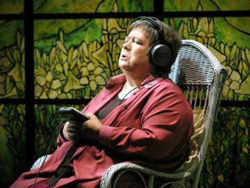 Punapukuinen vanhempi nainen istuu valkoisessa korituolissa, päässään suuret kuulokkeet ja kädessään kannettava audiosoitin, silmät kiinni, mahdollisesti kuunnellen musiikkia, lasimaalauksen edessä.