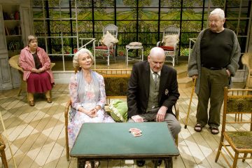 Neljä iäkästä ihmistä istuu ja seisoo pienen pöydän ympärillä pelikorttien kanssa valoisassa, viihtyisässä huoneessa, jossa on suuret ikkunat ja maalattu puutarhatausta. Tunnelma vaikuttaa mietiskelevältä ja epämuodolliselta.