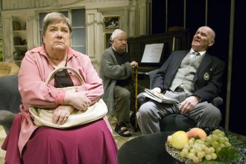 Kolme iäkästä ihmistä istuu olohuoneessa; vaaleanpunapukuinen nainen näyttää mietteliäältä, pukumies näyttää ahdistuneelta, ja toinen mies istuu pianon ääressä. Etualalla näkyy hedelmälautanen ja nuotteja.