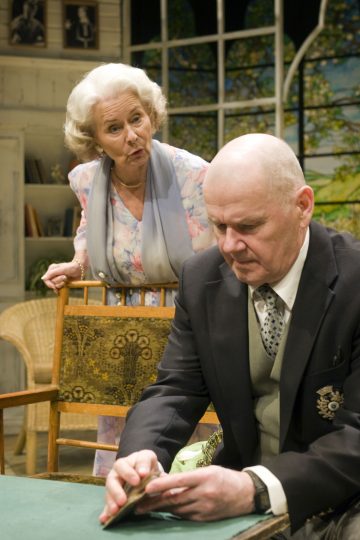 Valkohiuksinen iäkäs nainen seisoo pöydän ääressä istuvan vanhemman miehen takana. Nainen näyttää huolestuneelta, kun taas mies katsoo korttia kädessään. Ympäristö vaikuttaa kodikkaalta, kodikkaalta huoneelta, josta on näkymä puutarhaan.