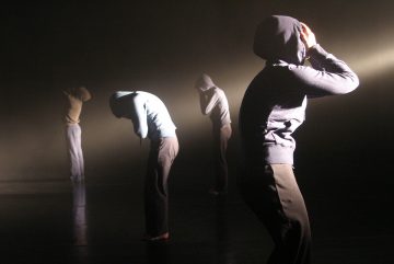 Neljä huppariin ja tummiin housuihin pukeutunutta ihmistä seisoo hämärässä, savuisessa tilassa, kukin pää kumartuneena ja kädet kasvoillaan, ja taustalta tuleva dramaattinen valonsäde valaisee heitä.