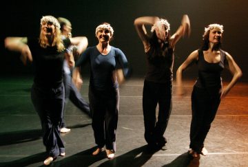 Neljä kukkakruunuihin ja tummiin vaatteisiin pukeutunutta henkilöä tanssii lavalla paljain jaloin ja liikuttaa käsiä energisesti. Tausta on hämärästi valaistu, mikä korostaa heidän iloisia ilmeitään ja dynaamisia asentojaan.