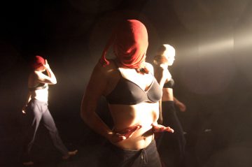 Kolme tummiin housuihin ja rintaliiveihin pukeutunutta ihmistä seisoo hämärässä tilassa, ja kullakin on päässään punainen kangas. Yksi seisoo edessä kädet vatsallaan, kaksi on taustalla; dramaattinen valaistus luo tunnelmallisen tunnelman.