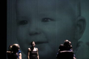 Suuri harmaasävykuva vauvan kasvoista heijastetaan näytölle. Etualalla seisoo useita aikuisia, joista jotkut syleilevät toisiaan, hämärässä valaistuksessa eri suuntiin päin.