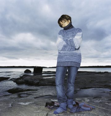 Siniseen villapaitaan ja farkkuihin pukeutunut henkilö seisoo veden äärellä kivikkoisella rannalla kädet ristissä lämpimänä. Huivi on löysästi kaulassa, ja taivas yllä on pilvinen ja pilvinen.