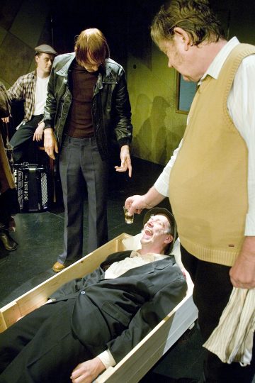 Neljä miestä esiintyy lavalla. Yksi mies makaa arkussa pukuun pukeutuneena, kun toinen kaataa juomaa suuhunsa. Kaksi muuta miestä katselee, toinen osoittaen, mikä luo dramaattisen ja intensiivisen kohtauksen.