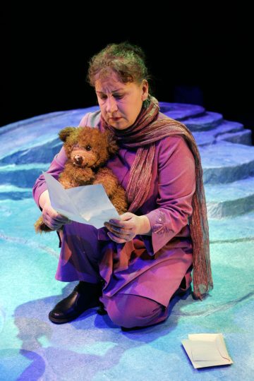 Sinisellä, kallioisella lavalla istuu violettiin asuun ja huiviin pukeutunut nainen, joka pitää nallea kädessään ja lukee paperia, jonka vieressä on toinen paperi maassa.