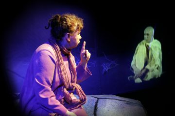 Violettiin asuun ja huiviin pukeutunut nainen istuu kalliolla, pitää sormeaan ylhäällä ikään kuin puhuisi, ja vastapäätä häntä istuu hehkuva, aavemainen hahmo pimeässä, teatterimaisessa ympäristössä, jossa on sininen ja violetti valaistus.