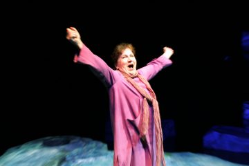 Vaaleanpunaiseen asuun ja huiviin pukeutunut nainen seisoo kädet kohotettuna ja suu auki iloisena tai voitonriemuisena tummalla taustalla, jossa on sinivihreitä muotoja.