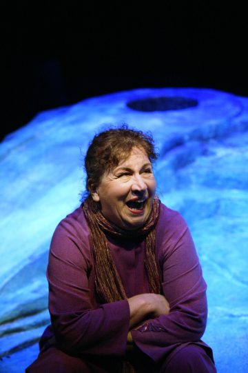 Ruskeakiharainen nainen, jolla on violetti toppi ja huivi, istuu iloisesti naureskellen. Hän on sinisellä valaistulla taustalla, joka muistuttaa kalliota tai abstraktia maisemaa.