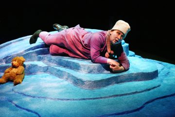Vaaleanpunaiseen asuun ja neulomyssyyn pukeutunut henkilö makaa sinisellä, jäätä muistuttavalla näyttämöllä nallen vieressä teatterivalaistuksessa.
