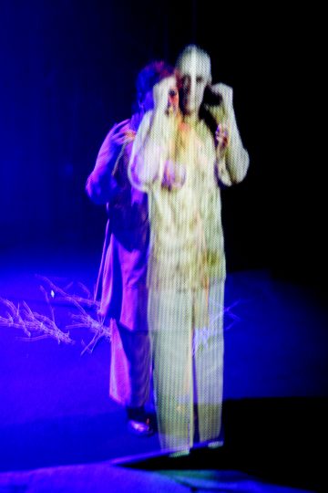 Kaksi ihmistä seisoo lähekkäin pimeällä näyttämöllä, jossa on sininen valaistus, ja toinen heistä on pukeutunut vaaleisiin vaatteisiin, jotka näyttävät puoliksi läpinäkyviltä, jolloin syntyy aavemainen tai kaksoisvalottunut vaikutelma. Lattialle on ripoteltu olkia tai oksia.