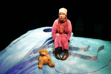 Vaaleanpunaisiin talvivaatteisiin pukeutunut henkilö istuu sinisellä, jäisellä lavalla ruskean nallen vieressä ja katsoo sivulle yllättyneenä. Tausta on tumma, mikä korostaa näyttämöä.