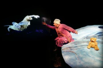 Vaaleanpunaiseen asuun ja valkoiseen hattuun pukeutunut henkilö istuu sinisellä, jäisellä näyttämöllä nallen vieressä ja kurottautuu kohti tummalla taustalla näkyvää uivan hahmon kuvaa.