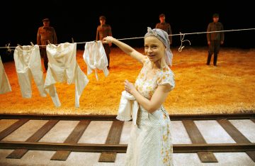 Kukkamekkoon ja huiviin pukeutunut nainen ripustaa valkoista pyykkiä junaradan yläpuolelle ripustettuun pyykkinaruun, ja taustalla seisoo neljä univormupukuista sotilasta olkipeitteisellä lavalla.