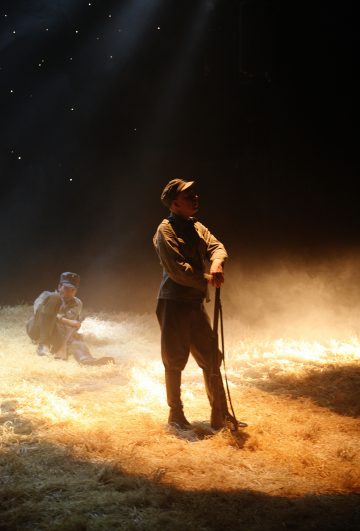 Lavalla on kaksi sotilaspukuista ihmistä dramaattisessa valaistuksessa; toinen seisoo kivääri kädessään ja toinen istuu maassa taustalla, molemmat olkien ja sumun ympäröimänä.