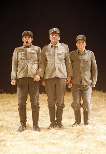 Kolme ihmistä seisoo rivissä olkipeitteisellä näyttämöllä, pukeutuneina samanlaisiin harmaisiin sotilasunivormuihin ja -lakkeihin, kasvot eteenpäin vakavat ja liioitellut ilmeet. Tausta on tumma.