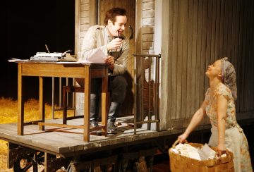 Mies istuu puisen kirjoituspöydän ääressä kirjoituskoneen kanssa, hymyilee ja puhuu pyykkikoria kantavan naisen kanssa. Kohtaus näyttää tapahtuvan kuistilla, jonka ympärille on ripoteltu heinää.