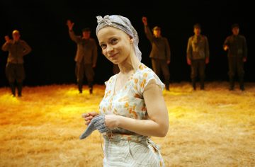 Kukkamekkoon ja huiviin pukeutunut nainen seisoo hymyillen olkipeitteisellä näyttämöllä neule kädessään, ja taustalla on neljä univormupukuista ihmistä.