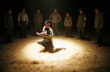 Sotilaspukuun pukeutunut henkilö polvistuu dramaattisesti oljen päällä valonheittimen alla, ja häntä ympäröi seitsemän muuta samanlaisiin univormuihin pukeutunutta henkilöä, jotka seisovat katsomassa pimeässä, teatraalisessa ympäristössä.