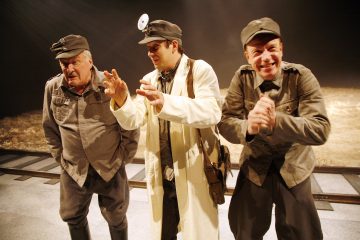 Lavalla esiintyy kolme pukumiestä; kahdella on harmaa sotilasunivormu ja lippis, kun taas keskellä olevalla on valkoinen takki, pääpeili ja salkku, ja hän vaikuttaa lääkäriltä. Tausta on hämärästi valaistu.