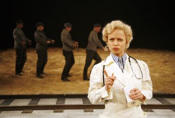 Vaaleahiuksinen nainen valkoisessa takissa pitelee muistivihkoa ja kynää ja vaikuttaa mietteliäältä. Neljä harmaapukuista miestä seisoo käsiraudoissa rivissä hänen takanaan lavalla, jossa on olkia ja junaratoja.