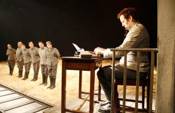 Etualalla mies kirjoittaa kirjoituspöydän ääressä, kun kuusi harmaisiin univormuihin pukeutunutta ihmistä seisoo rivissä oljilla päällystetyllä näyttämöllä taustalla, mikä viittaa dramaattiseen teatterikohtaukseen.