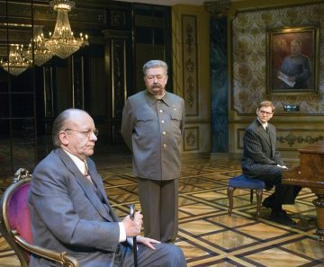 Kolme miestä on ylellisessä, koristeellisessa huoneessa. Yksi istuu keppi kädessään, toinen seisoo sotilaspuvussa ja kolmas istuu pianon ääressä. Heidän takanaan kuviotapetoidulla seinällä roikkuu suuri muotokuva univormupukuisesta miehestä.