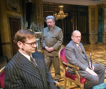 Kolme miestä ylellisessä, koristeellisesti koristellussa huoneessa: kaksi istuu punaisilla tuoleilla puvut yllään, kun taas yksi seisoo heidän takanaan sotilaspuvussa. Tunnelma vaikuttaa vakavalta tai jännittyneeltä.