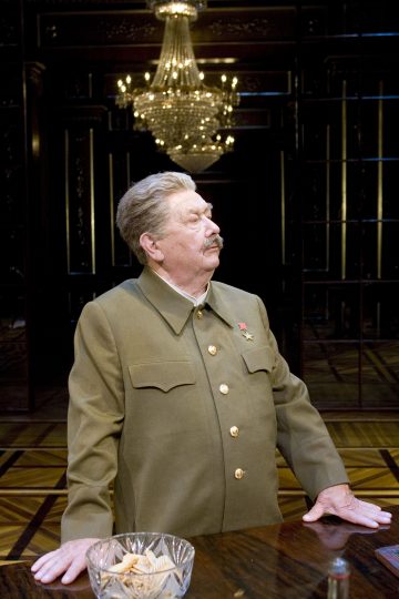 Vanhempi mies, jolla on harmaat hiukset ja viikset ja joka on pukeutunut vihreään sotilaspukuun, jossa on kultaiset napit ja mitali, seisoo pöydän vieressä koristeellisessa huoneessa, jossa on kattokruunu.