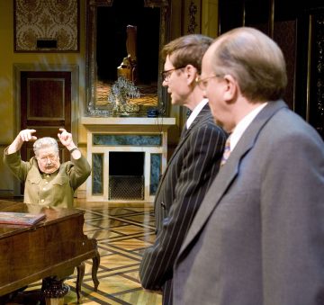 Vanhempi nainen elehtii eloisasti pianon ääressä istuen, kun hän on vastapäätä kahta pukumiestä, jotka seisovat selkä kameraan päin koristeellisesti sisustetussa huoneessa.