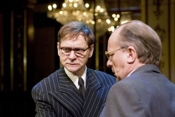 Kaksi pukumiestä seisoo sisätiloissa kattokruunun alla. Toinen mies, jolla on silmälasit ja raidallinen puku, näyttää vakavalta ja katsoo hieman kameraan päin, kun taas toinen mies katsoo poispäin ja näyttää vain profiilinsa.