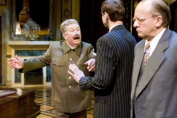 Vanhempi mies sotilaspuvussa osoittaa vihaisesti kahdelle pukumiehelle, joista toinen on häntä vastapäätä ja toinen seisoo oikealla varautunut ilme kasvoillaan koristeellisessa, hyvin valaistussa huoneessa.