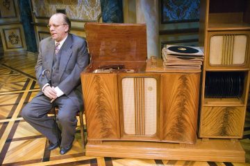 Vanhempi pukumies istuu puisella tuolilla keppi kädessään vanhan puisen levysoittimen vieressä, jonka taustalla on pinottuja vinyylilevyjä ja koristeellinen sisustus.