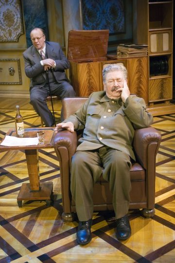 Kaksi miestä koristeellisesti sisustetussa huoneessa; toinen, vihreään sotilasunivormuun pukeutunut, istuu ruskealla nojatuolilla mietteliään näköisenä, kun taas toinen, pukuun pukeutunut ja keppi kädessään, istuu hänen takanaan. Nojatuolin vieressä on pöytä, jossa on pullo ja lasi.