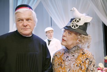 Musta pappispukuinen vanhempi mies seisoo vanhemman naisen vieressä, jolla on koristeellinen hattu, jonka päällä on väärennetty valkoinen kyyhkynen ja pyörä; taustalla on kolmas henkilö valkoisessa univormussa.