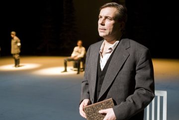 Etualalla seisoo pukumies, joka pitää kirjaa kädessään ja näyttää mietteliäältä. Taustalla on kaksi muuta miestä hämärässä, toinen istumassa ja toinen seisomassa, heikosti valaistulla näyttämöllä.