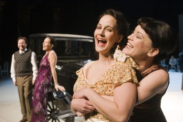 Etualalla kaksi vintage-mekkoon pukeutunutta naista nauraa ja syleilee, kun taas taustalla mies ja nainen seisovat hymyillen vanhanaikaisen auton vieressä. Kohtaus näyttää olevan teatteriesityksestä.